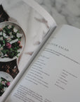 Open cookbook showing a salad recipe with a bowl of salad on a marble surface