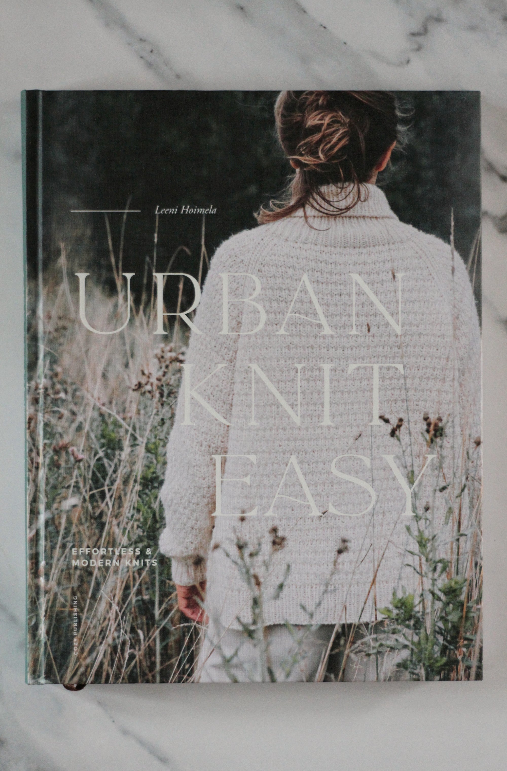 Book titled 'The Urban Knit Easy' with a person wearing a knitted garment on a marble surface.